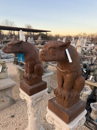 Two brown gargoyle statues perched on ornate white pedestals at an outdoor garden store.
