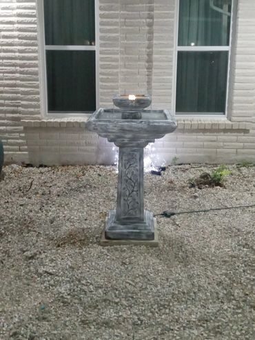 Stone birdbath fountain in a gravel garden in front of a brick house.