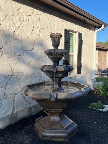 A three-tiered stone water fountain outside a house with a stone wall.