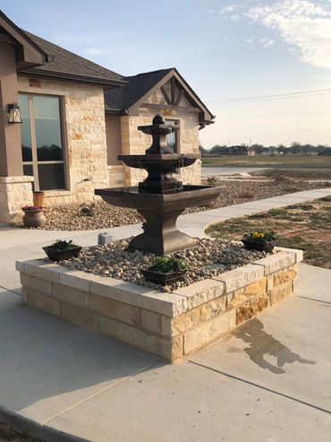 Stone water fountain in front of a beige house with rocky landscaping.