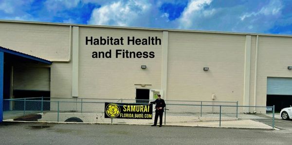 Building with Habitat Health and Fitness sign and a man standing by a Samurai Florida Budo banner.