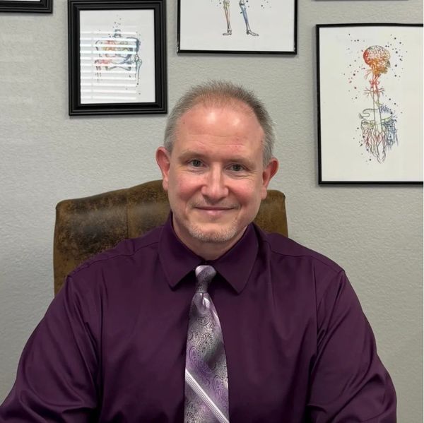 Man in purple shirt and tie sitting in an office with anatomy art on the wall.