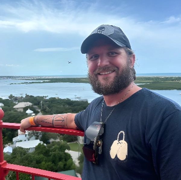 Smiling man with a beard standing on a red balcony overlooking water and greenery.
