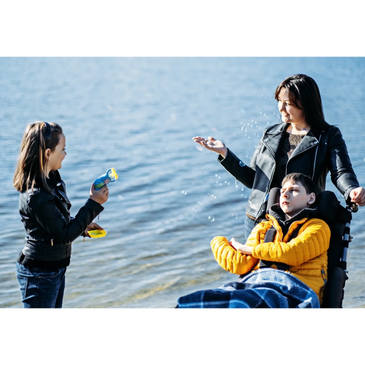 A young woman and kids at the beach are playing. A teen boy in a wheelchair and girl with bubble gun