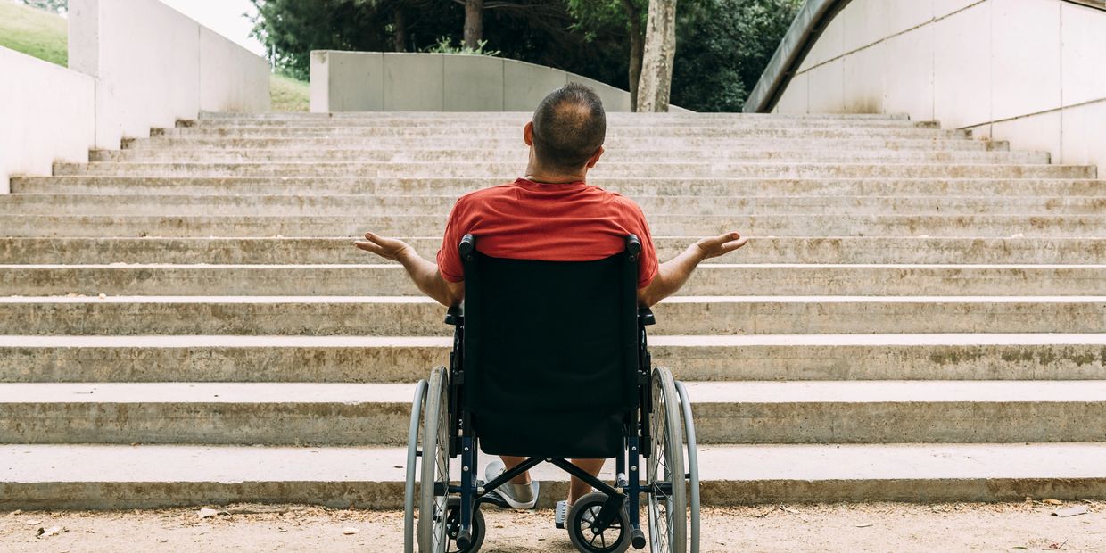 A frustrated man in a wheelchair is at the bottom of a lot of steps, Access Modification would help.