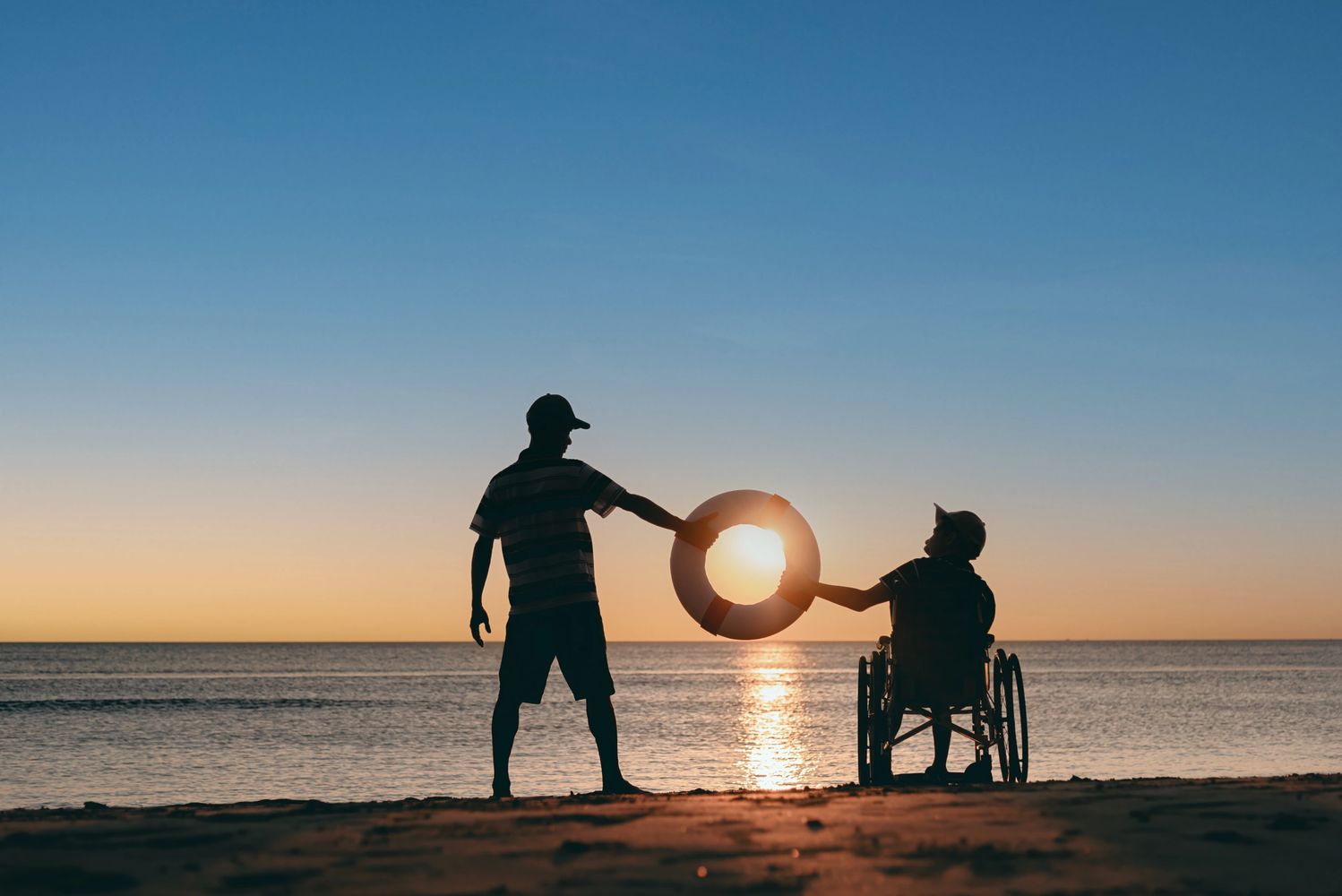 A wheelchair user and a friend are watching a sunset on the beach, hold a life ring framing the sun.