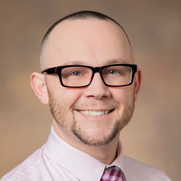 Smiling man with glasses wearing a pink shirt and plaid tie.