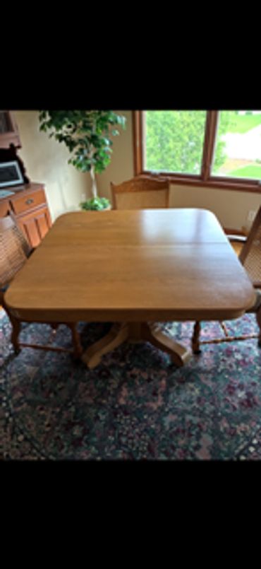 Wooden dining table with four chairs on a floral carpet near a window.