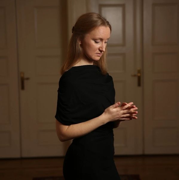 Woman in black dress looking down with folded hands indoors.