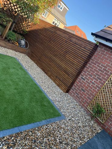 A neat backyard with artificial grass, gravel, and wooden fencing under a clear blue sky.