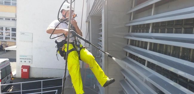 Worker in safety gear power washing a building facade while suspended.