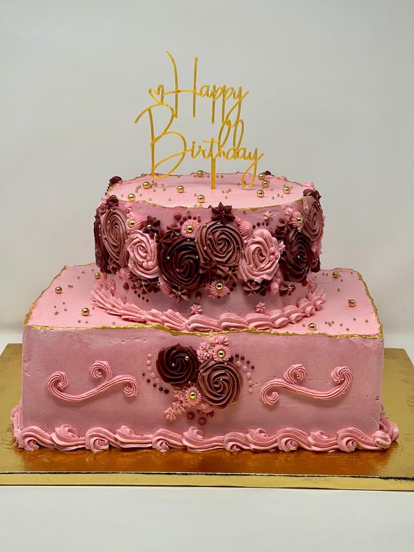 Pink 2-tier cake with 3 colored rosettes on the side of the top tier with a gold cake topper saying 