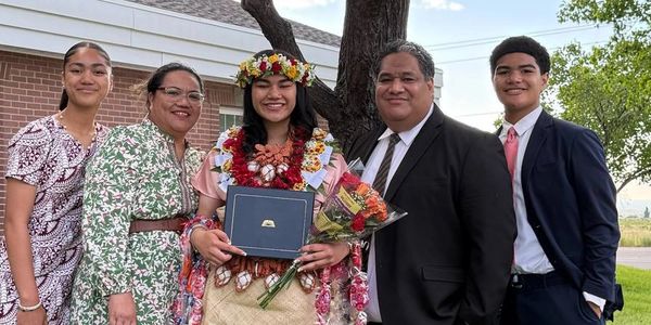 Family celebrates a young woman's graduation outdoors by a tree.