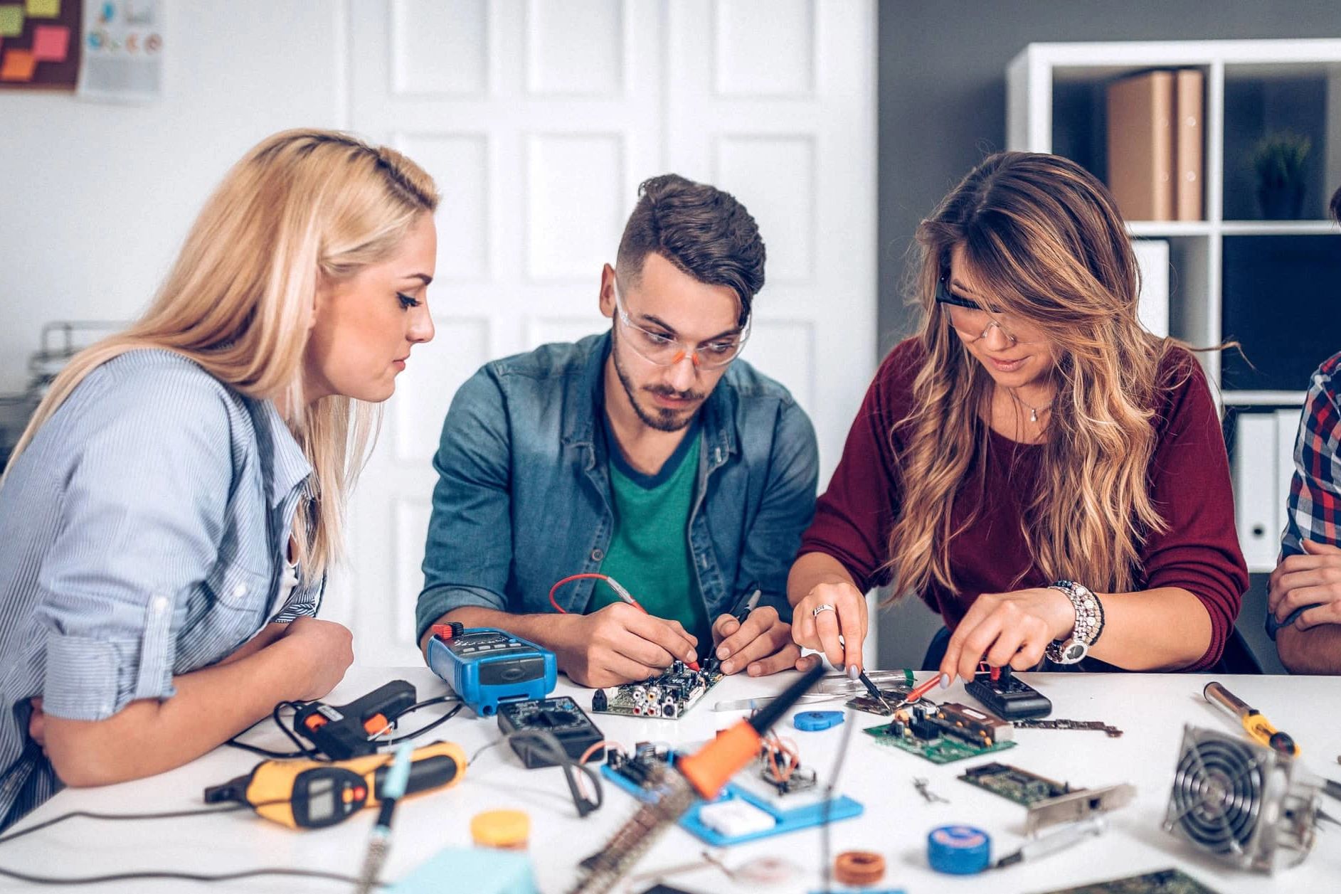 Three young people working intently on electronics projects at a cluttered table.