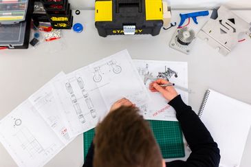 Person working on mechanical drawings at a desk with tools and parts.