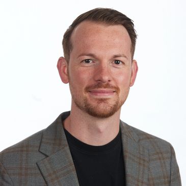 Man in a plaid blazer and black shirt smiling against a white background.