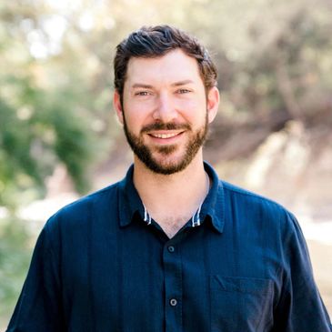 Smiling man with beard wearing a dark blue shirt outdoors.