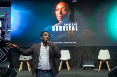Man speaking on stage with a 'Shades of Survival' poster behind.