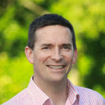 Man in a pink striped shirt smiling outdoors.
