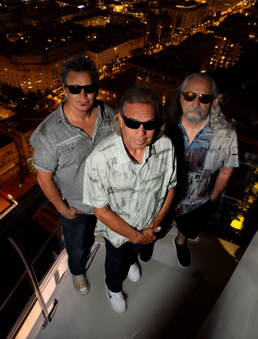 Three men wearing sunglasses pose on a rooftop at night.