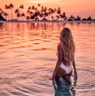 Woman in white swimsuit wading into water during sunset with palm trees in the background.