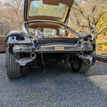 A stripped-down car missing rear parts, parked outdoors on gravel.