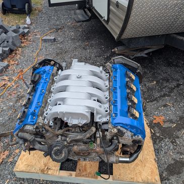 A car engine with blue valve covers and a silver intake manifold on a wooden platform outdoors.