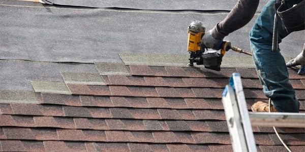 Worker installing asphalt shingles on a roof using a nail gun.