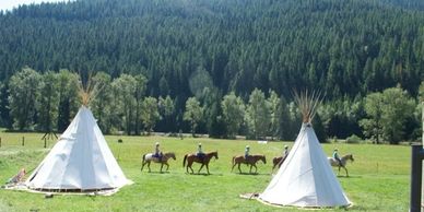 Two tipis on a green field with people riding horses in the background.