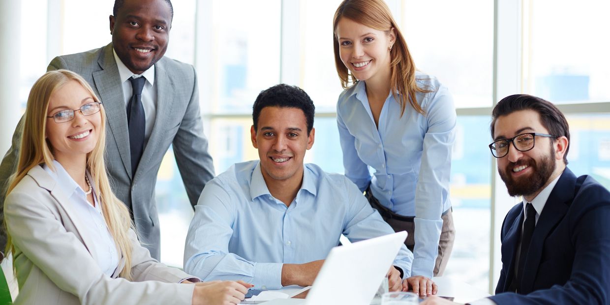 A diverse group of professionals smiling around a laptop in an office.