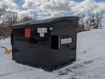 A black dumpster on a snowy ground under a cloudy sky.
