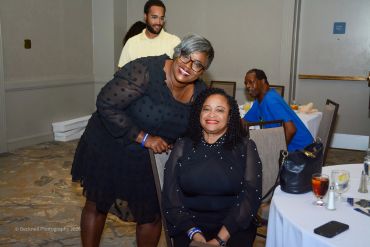 Two women smiling at a formal event, one standing and one seated.