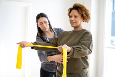 A woman exercises with a resistance band guided by a trainer indoors.