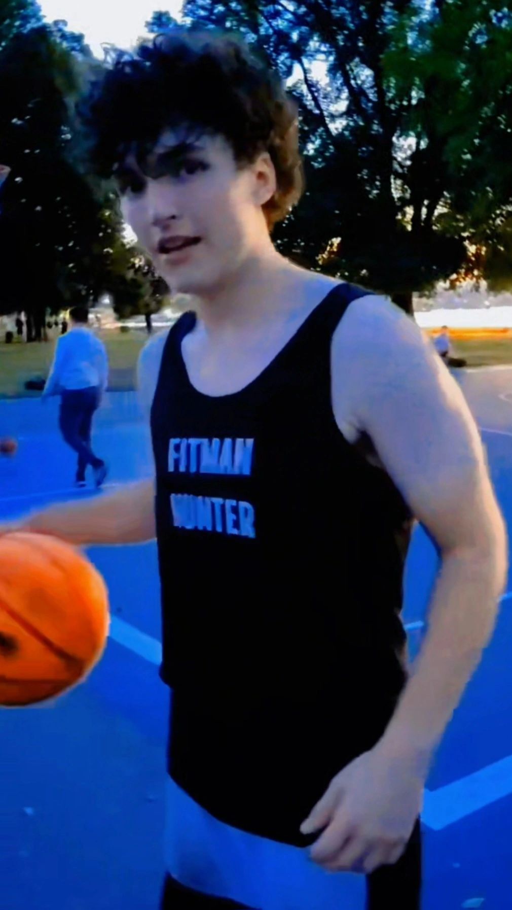 Young man holding a basketball on an outdoor court at dusk.