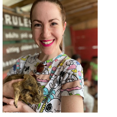 White femme with her hair up, smiling, wearing colorful shirt and holding a guinea pig in her arms