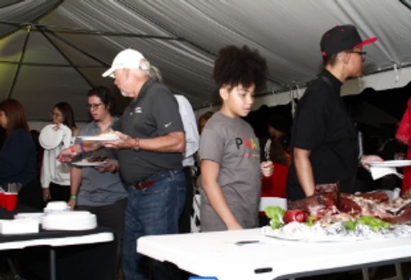 People serving and eating food at a buffet-style event under a tent.