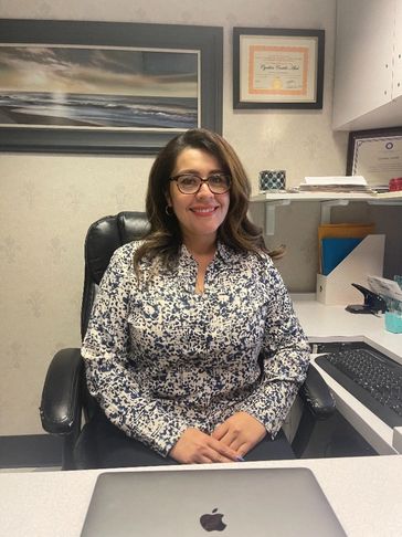 Woman sitting at an office desk, smiling with glasses and a patterned blouse.