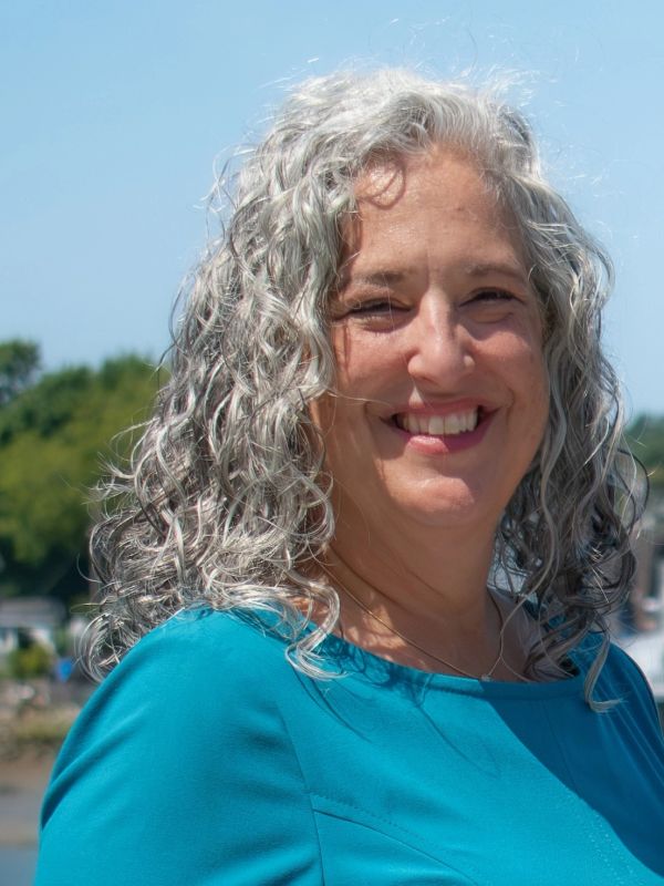 Smiling woman with curly gray hair wearing a blue top outdoors.