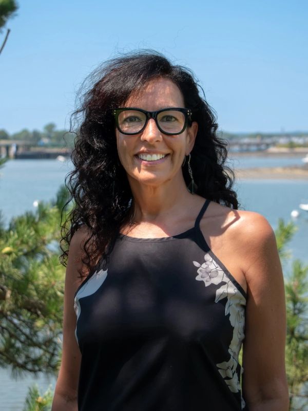 Smiling woman with curly hair and glasses near a waterfront.