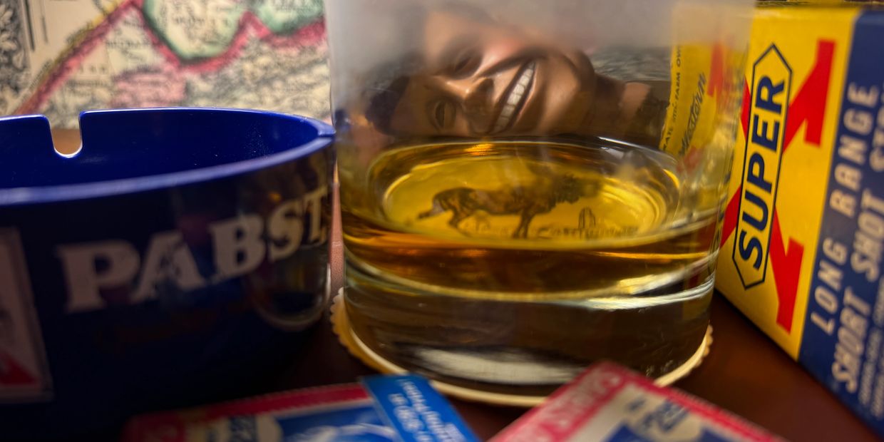 Whiskey glass reflecting a smiling face, with an ashtray and Super-X box.