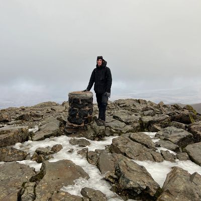 Person standing on rocky terrain beside a trig point under cloudy sky.
