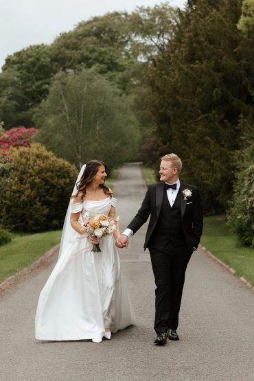 Newlyweds walk hand in hand down a tree-lined path, smiling joyfully.