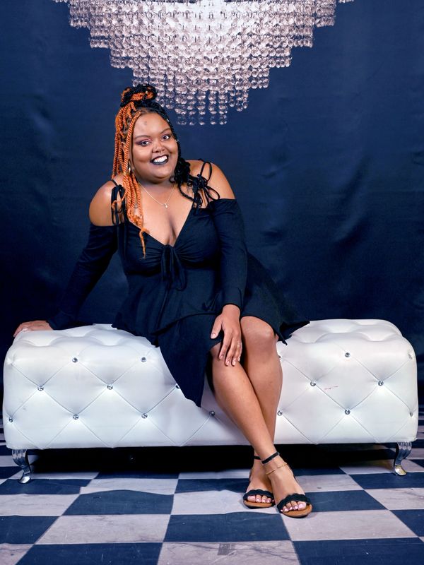 A woman with orange braids in a black dress sitting on a white tufted bench.