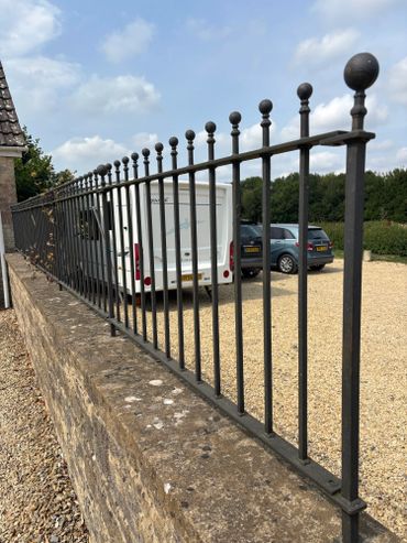Black metal fence on a stone wall with parked vehicles behind it.