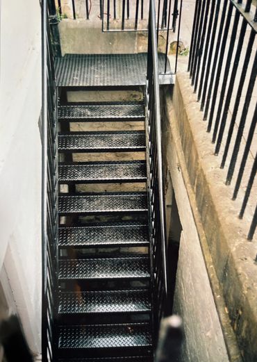 A narrow metal staircase descending between two walls with black railings.