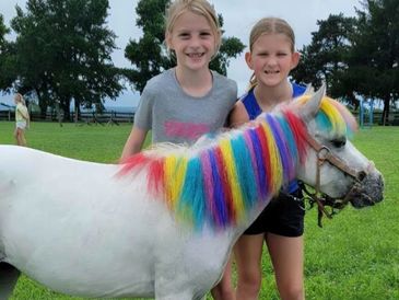 Two girls smiling next to a white pony with a rainbow-colored mane and tail.