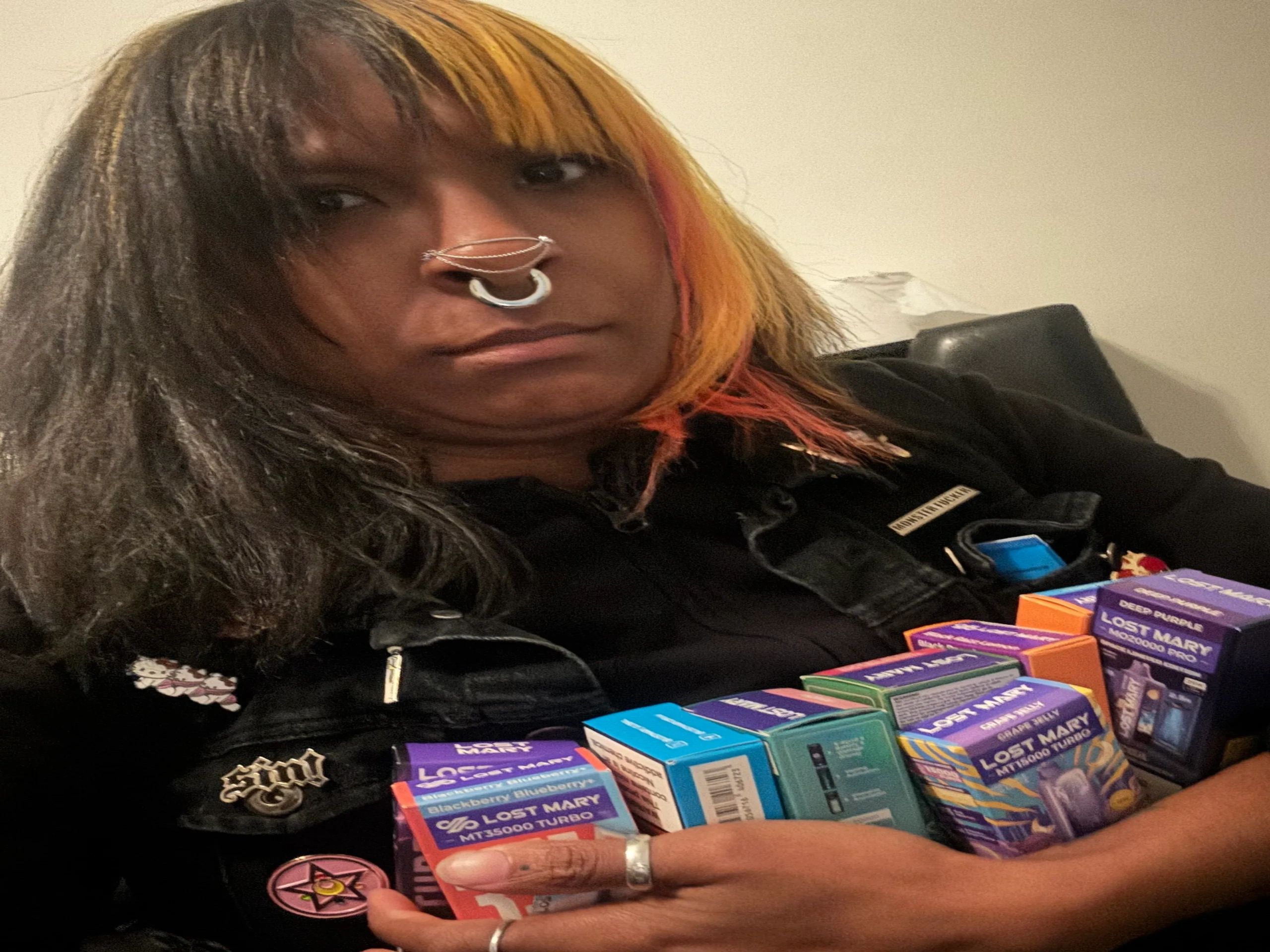 Person with septum piercing and multicolored hair holding vape boxes.