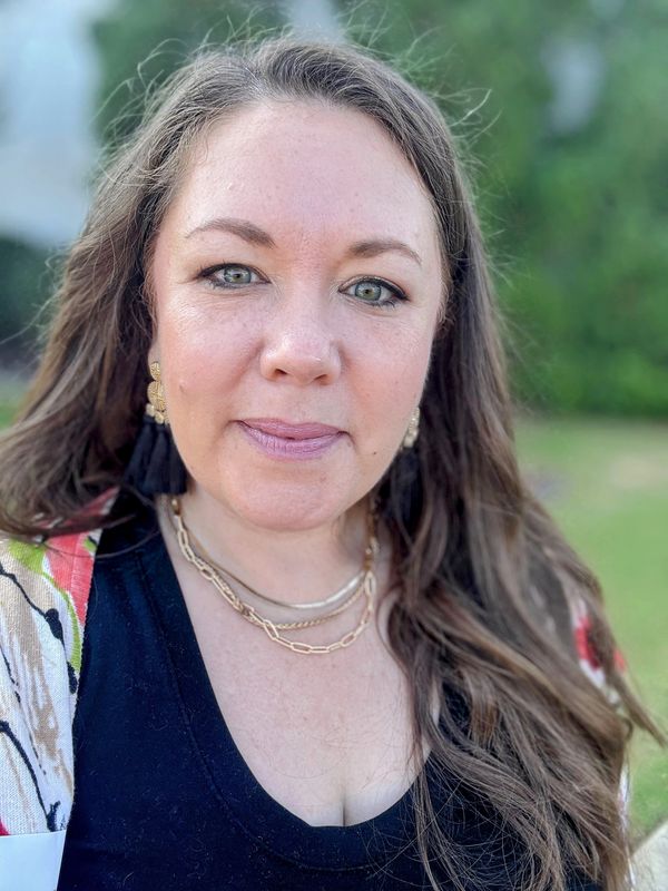 Woman with long brown hair outdoors, wearing gold jewelry.