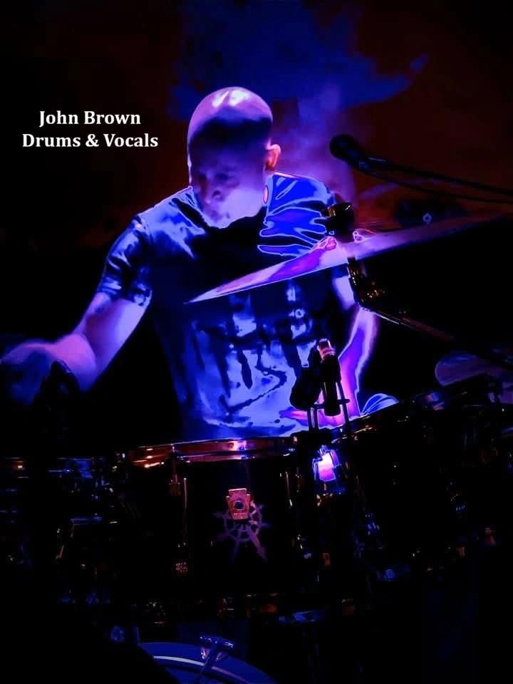 John Brown playing drums and singing on stage with blue lighting.