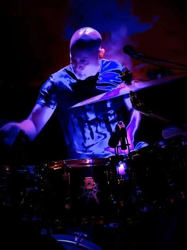 A drummer illuminated by blue and purple stage lights playing drums intensely.